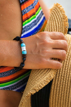 A turquoise and white beaded bracelet rests across the brim of a straw sunhat, close-up beach style.