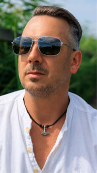 A man in sunglasses wears a leather necklace with a Thors hammer pendant, outdoors in natural light.