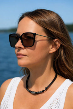 A woman in sunglasses wears a leather zodiac bead necklace, shown in profile against a lake backdrop.